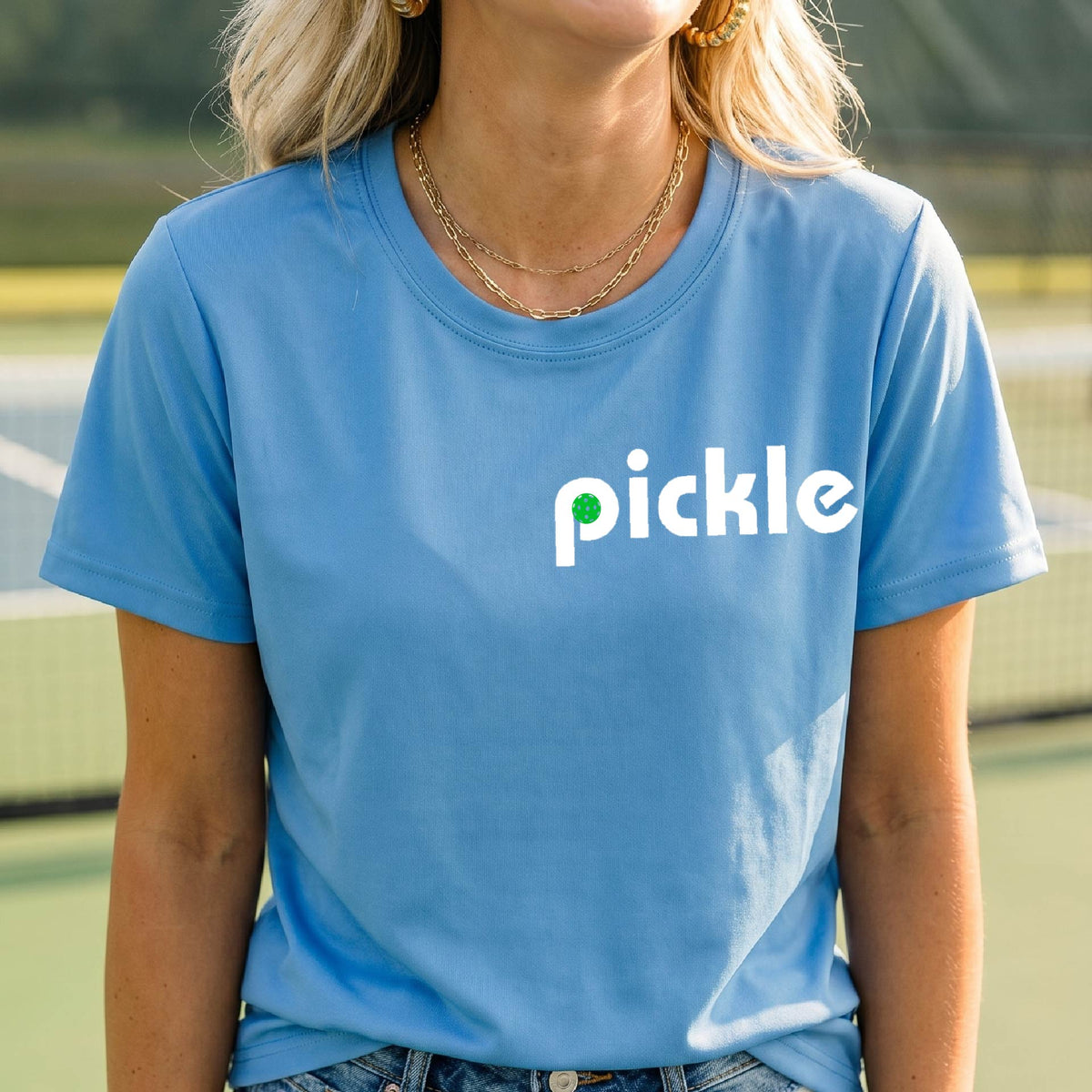 A woman wearing a blue short-sleeve t-shirt with a white graphic of ‘PICKLE’ with a green pickleball integrated into the letter P printed on the front, in front of a pickleball court. 