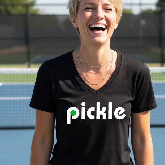 A woman wearing a black short-sleeve v-neck t-shirt with a white graphic of ‘PICKLE’ with a green pickleball integrated into the letter P printed on the front, in front of a pickleball court. 