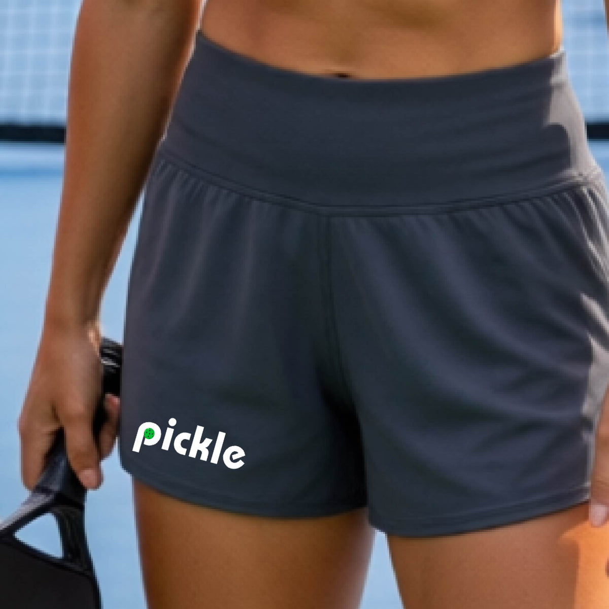 A close-up of a person wearing dark gray athletic shorts with the word 'pickle' printed in white and green on the left leg, on a blue pickleball court.