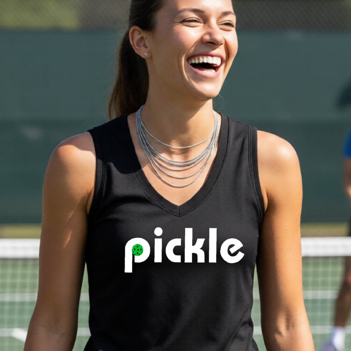 A woman wearing a black sleeveless v-neck t-shirt with a white graphic of ‘PICKLE’ with a green pickleball integrated into the letter P printed on the front, in front of a pickleball court. 