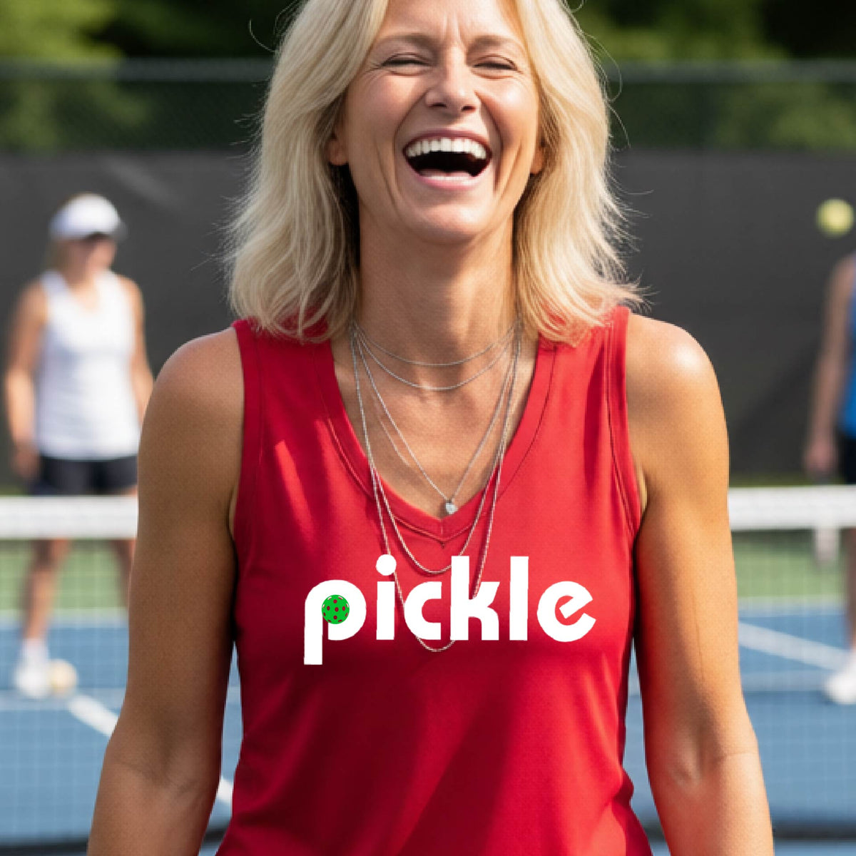 A woman wearing a red sleeveless v-neck t-shirt with a white graphic of ‘PICKLE’ with a green pickleball integrated into the letter P printed on the front, in front of a pickleball court. 