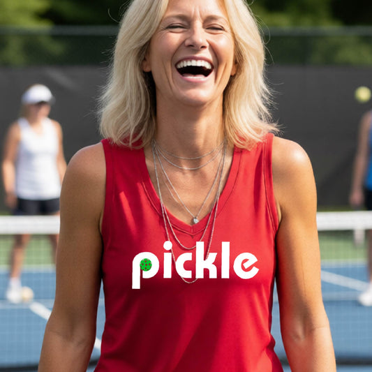 A woman wearing a red sleeveless v-neck t-shirt with a white graphic of ‘PICKLE’ with a green pickleball integrated into the letter P printed on the front, in front of a pickleball court. 