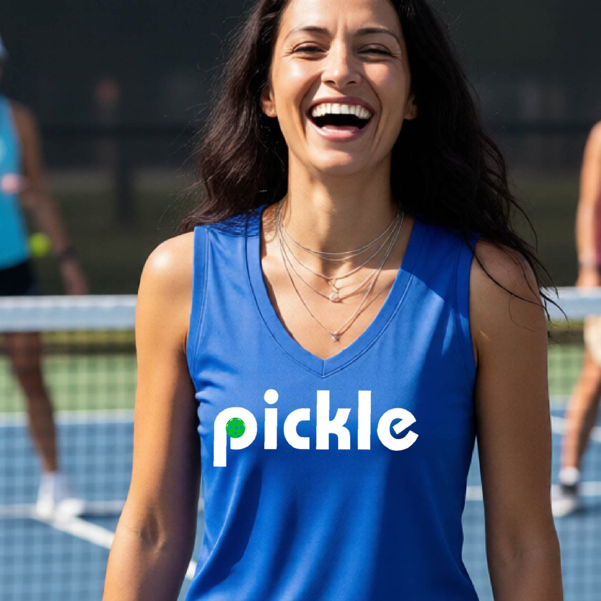 A woman wearing a royal blue sleeveless v-neck t-shirt with a white graphic of ‘PICKLE’ with a green pickleball integrated into the letter P printed on the front, in front of a pickleball court. 