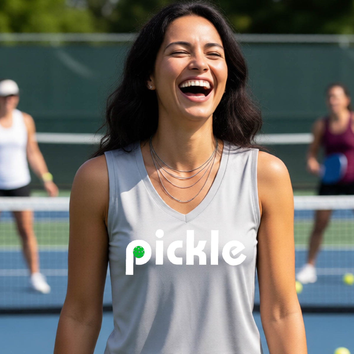 A woman wearing a silver gray sleeveless v-neck t-shirt with a white graphic of ‘PICKLE’ with a green pickleball integrated into the letter P printed on the front, in front of a pickleball court. 