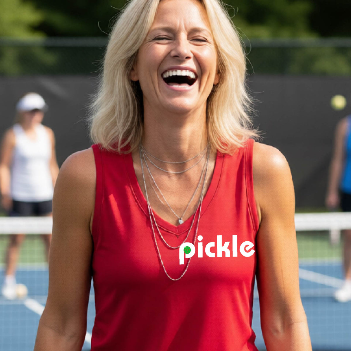 A woman wearing a red sleeveless v-neck t-shirt with a white graphic of ‘PICKLE’ with a green pickleball integrated into the letter P printed on the front, in front of a pickleball court. 