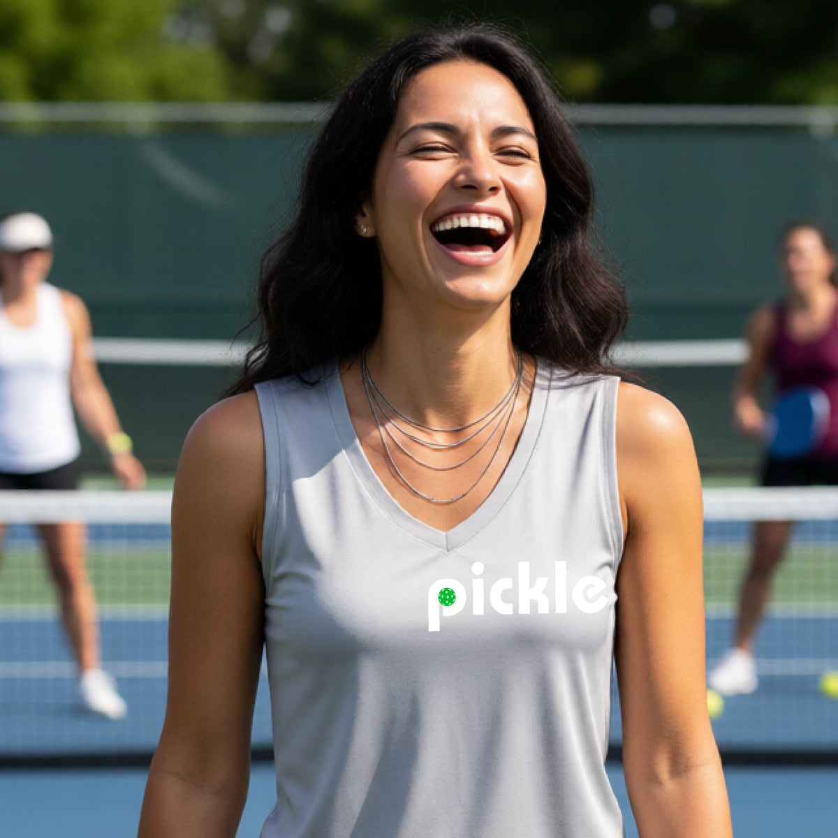A woman wearing a silver gray sleeveless v-neck t-shirt with a white graphic of ‘PICKLE’ with a green pickleball integrated into the letter P printed on the front, in front of a pickleball court. 