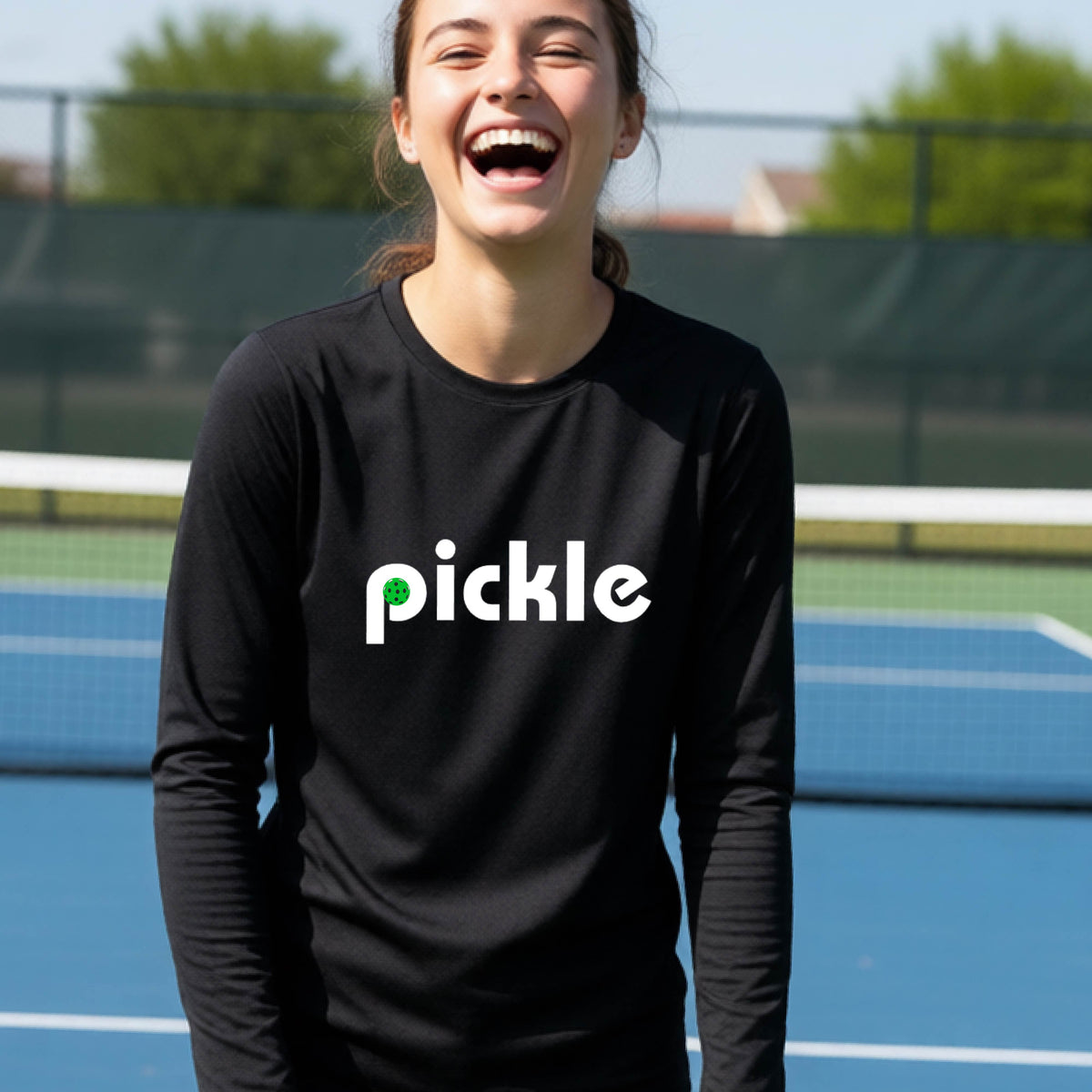 A young person laughing on a pickleball court wearing a black long-sleeve shirt with the word 'pickle' and a pickleball integrated into the P printed across the chest.