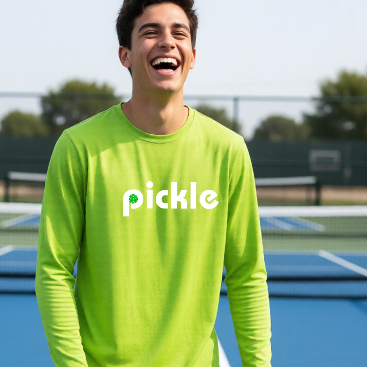 A young person laughing on a pickleball court wearing a lime green long-sleeve shirt with the word 'pickle' and a pickleball integrated into the P printed across the chest.