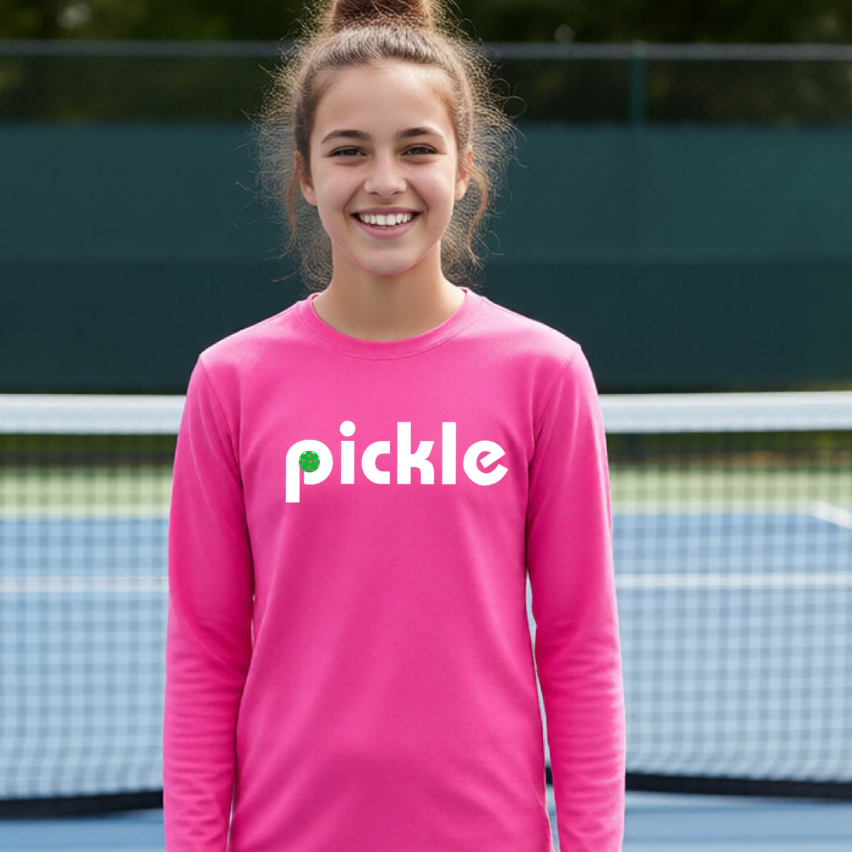 A young person laughing on a pickleball court wearing a pinklong-sleeve shirt with the word 'pickle' and a pickleball integrated into the P printed across the chest.