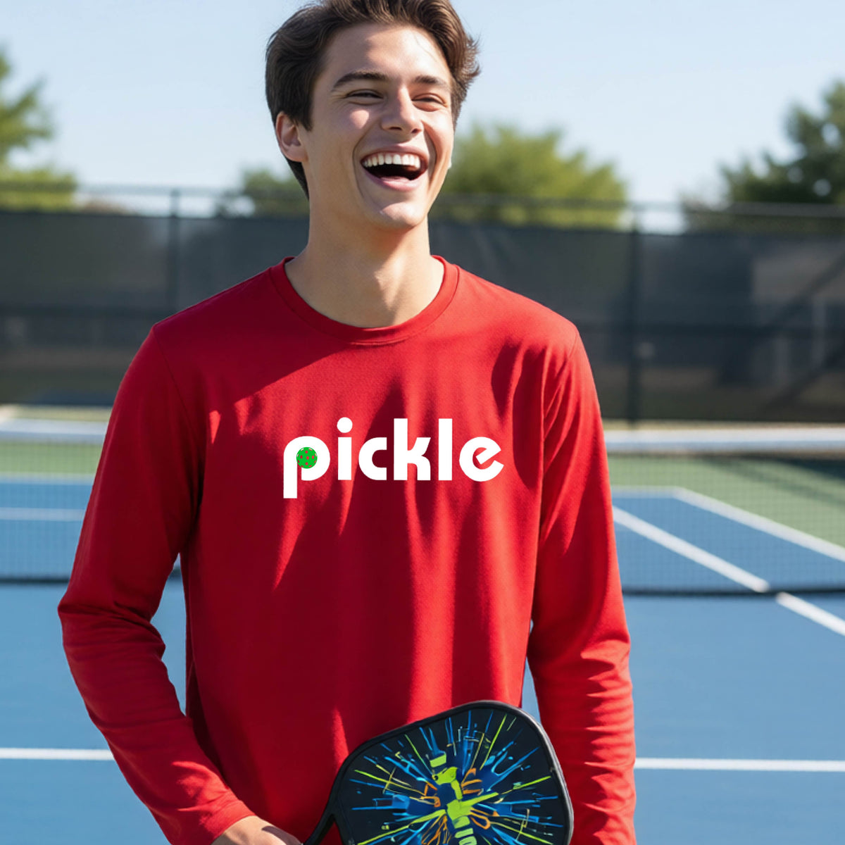 A young person laughing on a pickleball court wearing a red long-sleeve shirt with the word 'pickle' and a pickleball integrated into the P printed across the chest.