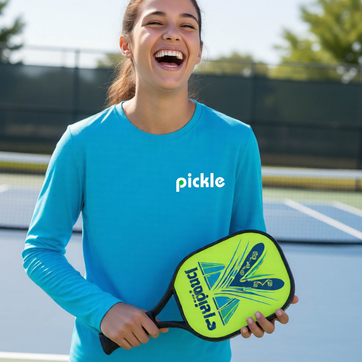 A young person laughing on a pickleball court wearing a bright blue long-sleeve shirt with the word 'pickle' and a pickleball integrated into the P printed across the chest.