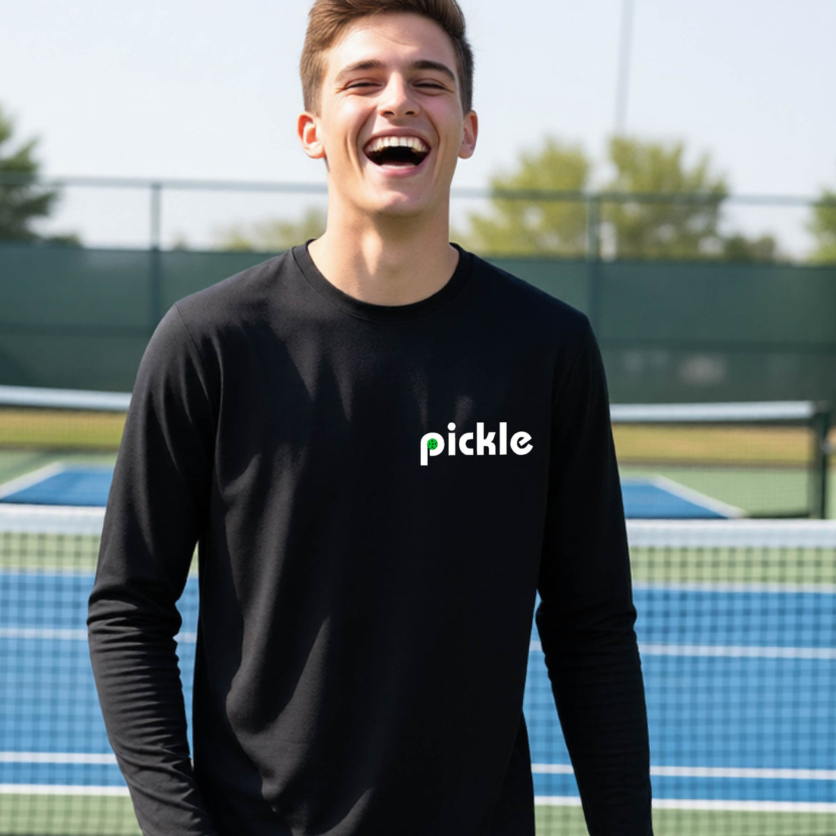 A young person laughing on a pickleball court wearing a black long-sleeve shirt with the word 'pickle' and a pickleball integrated into the P printed across the chest.