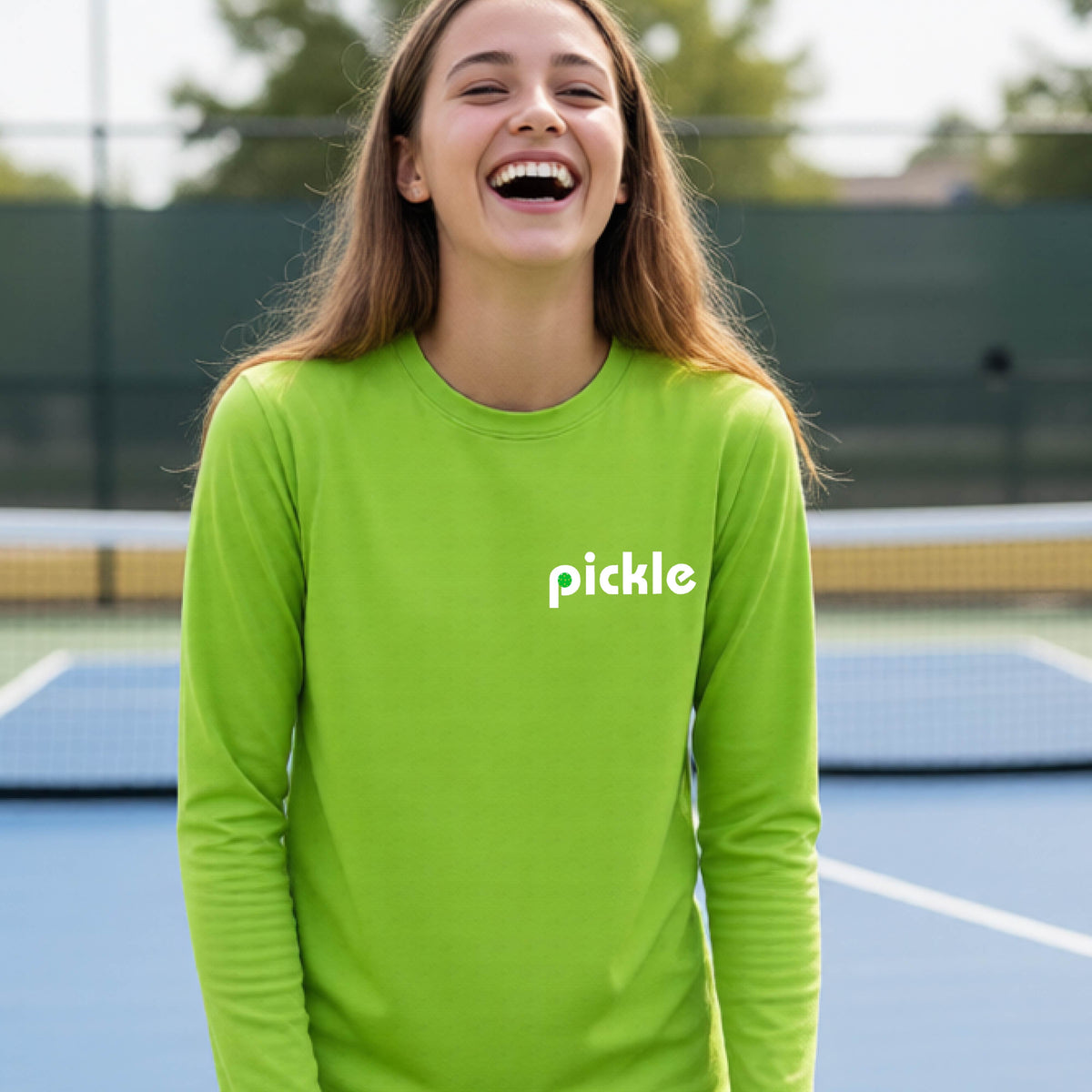 A young person laughing on a pickleball court wearing a lime green long-sleeve shirt with the word 'pickle' and a pickleball integrated into the P printed across the chest.