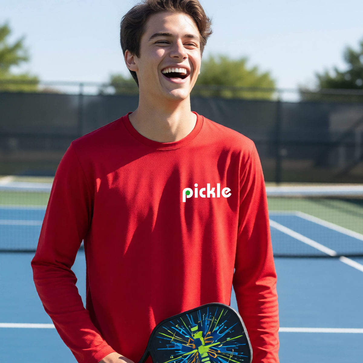 A young person laughing on a pickleball court wearing a red long-sleeve shirt with the word 'pickle' and a pickleball integrated into the P printed across the chest.