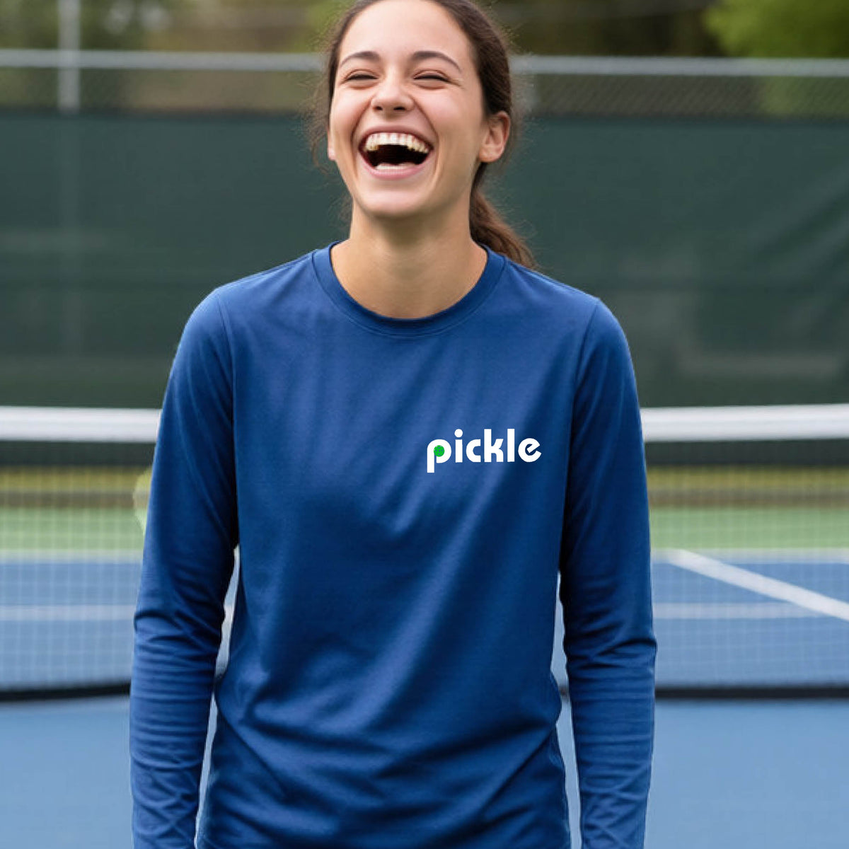 A young person laughing on a pickleball court wearing a royal blue long-sleeve shirt with the word 'pickle' and a pickleball integrated into the P printed across the chest.