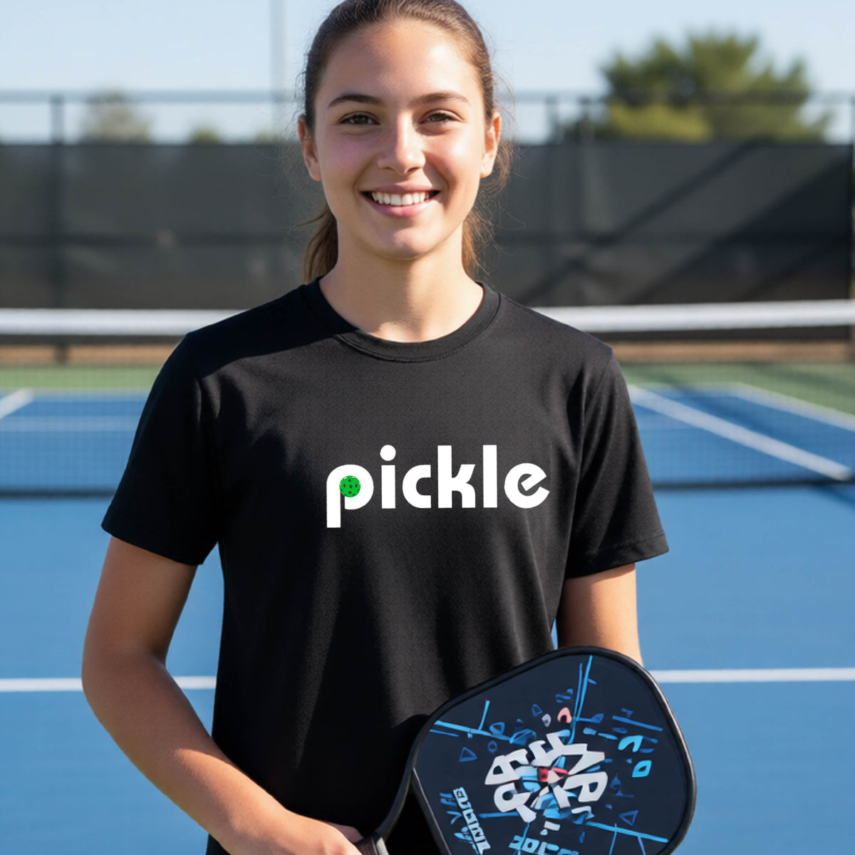 A young person wearing a black short-sleeve t-shirt with a white graphic of ‘PICKLE’ with a green pickleball integrated into the letter P printed on the front, in front of a pickleball court. 