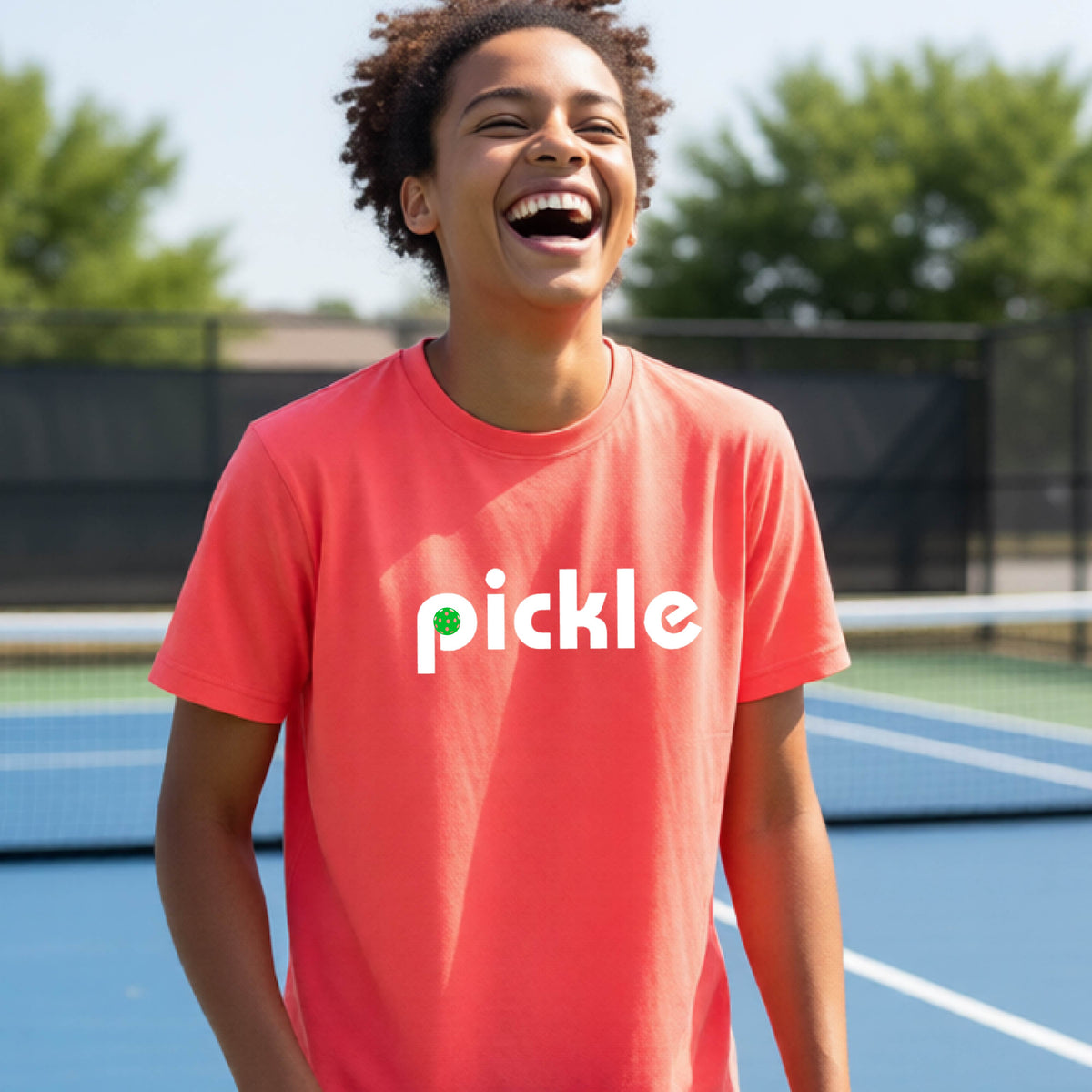 A young person wearing a coral pink short-sleeve t-shirt with a white graphic of ‘PICKLE’ with a green pickleball integrated into the letter P printed on the front, in front of a pickleball court. 