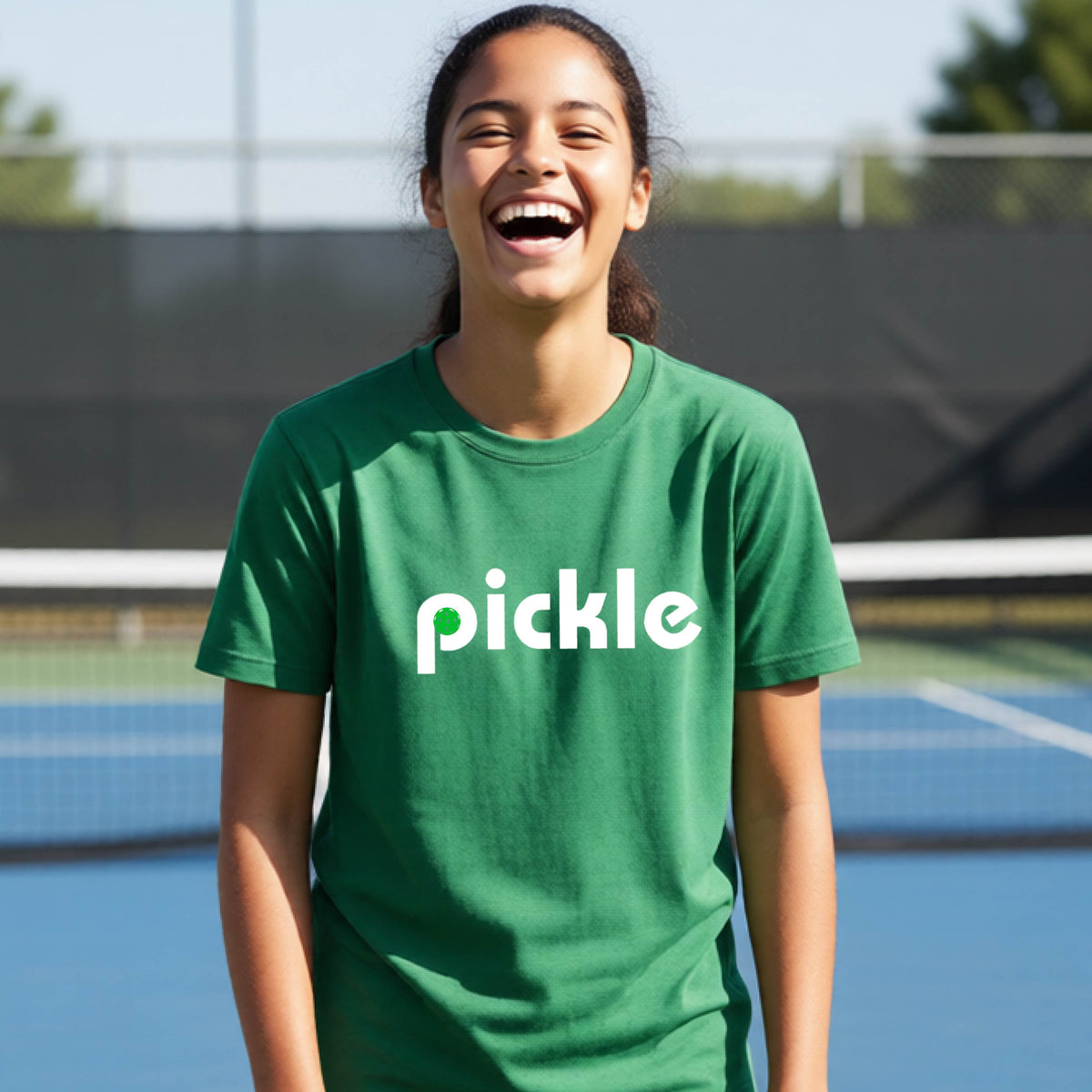 A young person wearing a green short-sleeve t-shirt with a white graphic of ‘PICKLE’ with a green pickleball integrated into the letter P printed on the front, in front of a pickleball court. 