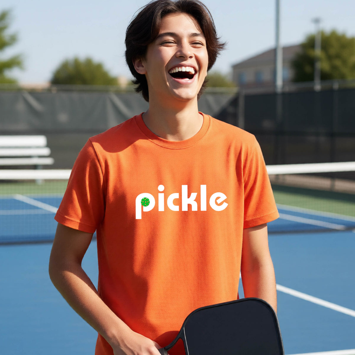 A young person wearing an orange short-sleeve t-shirt with a white graphic of ‘PICKLE’ with a green pickleball integrated into the letter P printed on the front, in front of a pickleball court. 