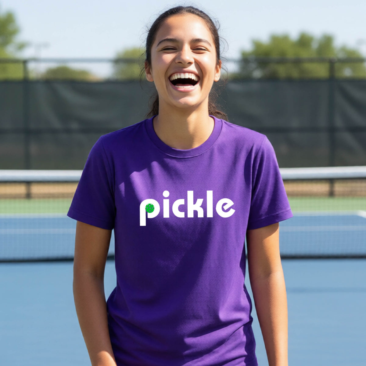 A young person wearing a purple short-sleeve t-shirt with a white graphic of ‘PICKLE’ with a green pickleball integrated into the letter P printed on the front, in front of a pickleball court. 