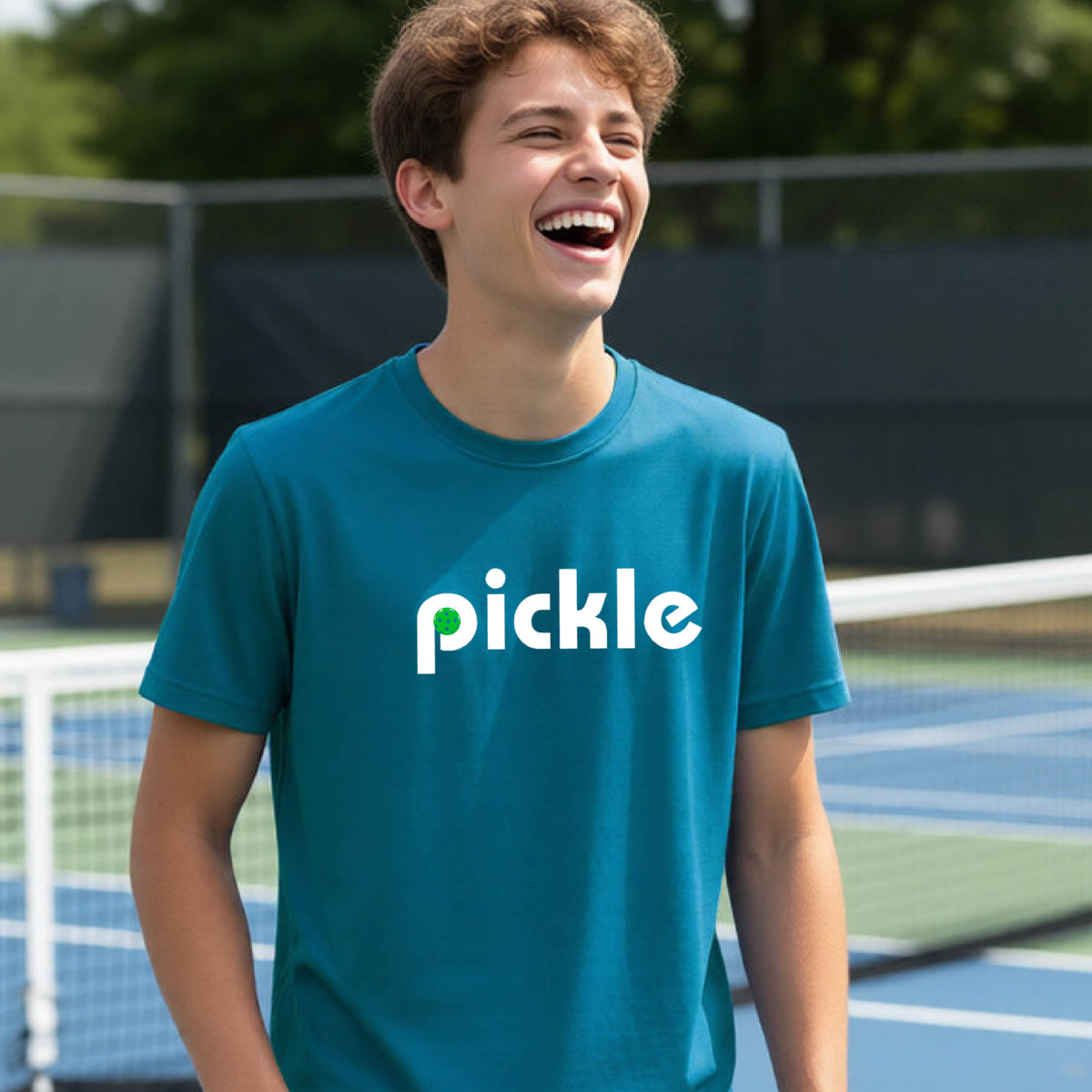 A young person wearing a teal short-sleeve t-shirt with a white graphic of ‘PICKLE’ with a green pickleball integrated into the letter P printed on the front, in front of a pickleball court. 
