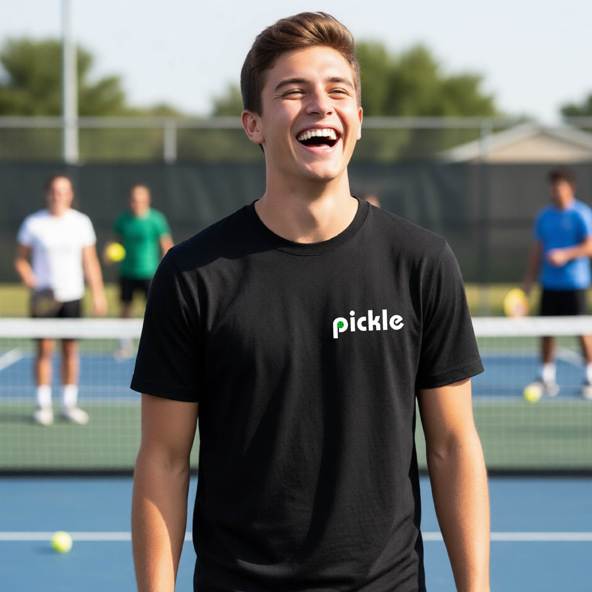 A young person wearing a black short-sleeve t-shirt with a white graphic of ‘PICKLE’ with a green pickleball integrated into the letter P printed on the front, in front of a pickleball court. 