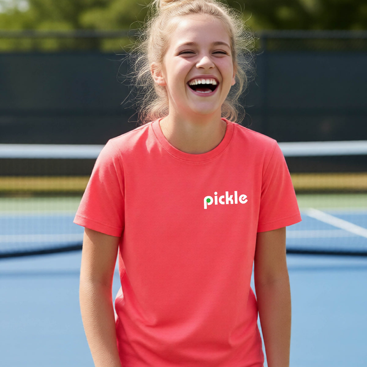 A young person wearing a coral pink short-sleeve t-shirt with a white graphic of ‘PICKLE’ with a green pickleball integrated into the letter P printed on the front, in front of a pickleball court. 