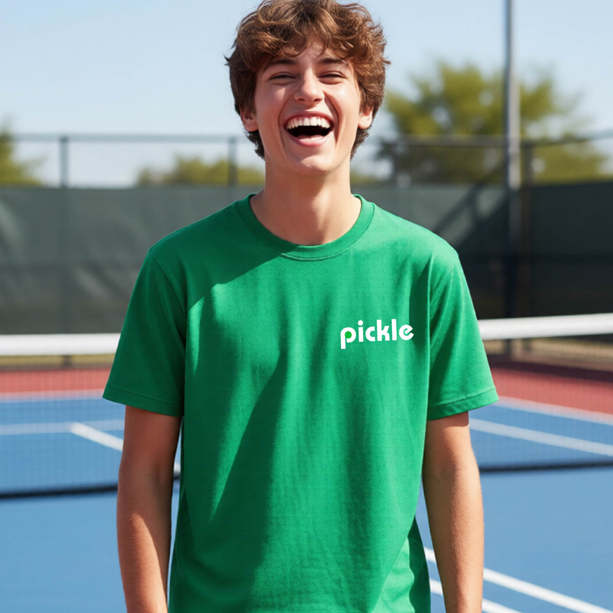 A young person wearing a green short-sleeve t-shirt with a white graphic of ‘PICKLE’ with a green pickleball integrated into the letter P printed on the front, in front of a pickleball court. 