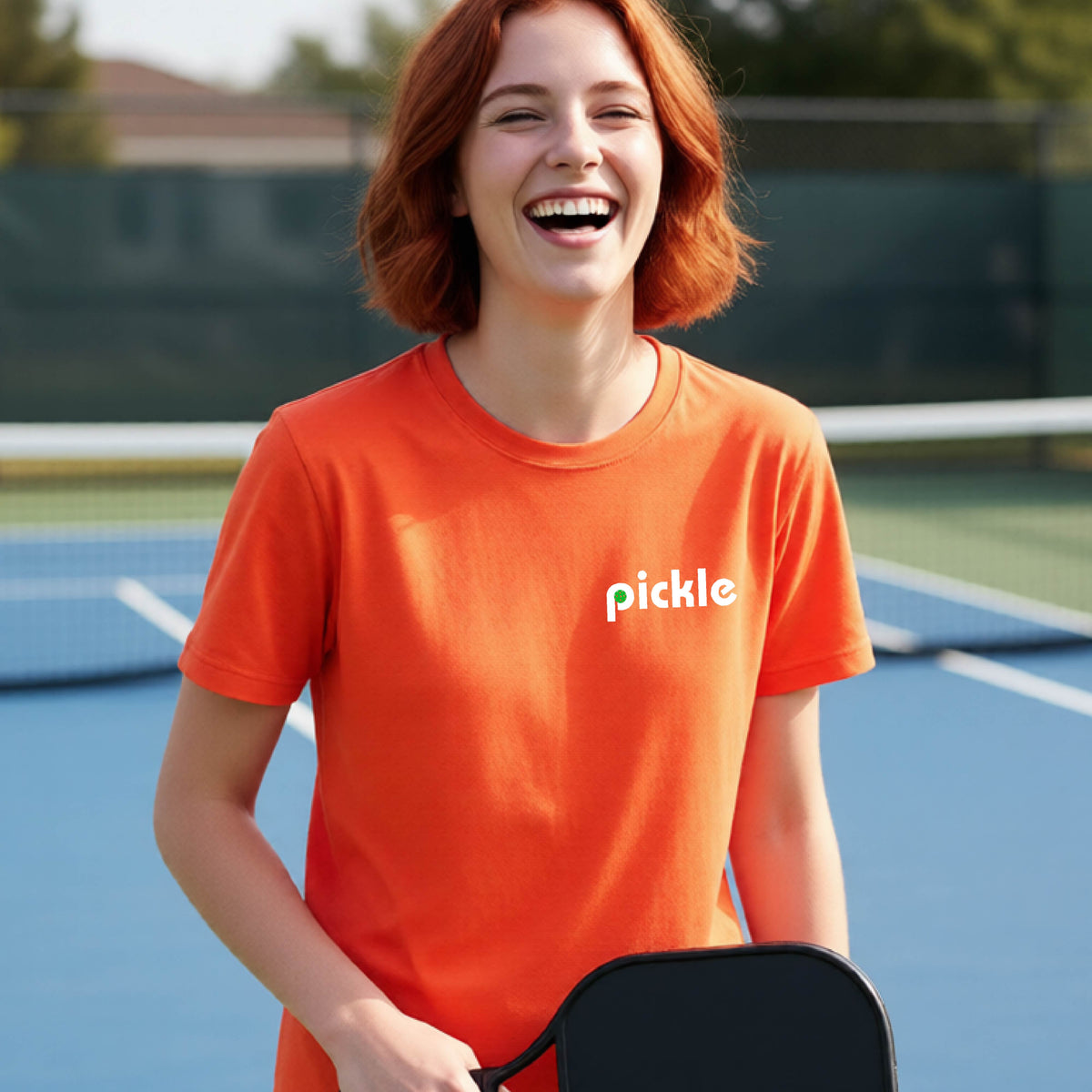 A young person wearing an orange short-sleeve t-shirt with a white graphic of ‘PICKLE’ with a green pickleball integrated into the letter P printed on the front, in front of a pickleball court. 