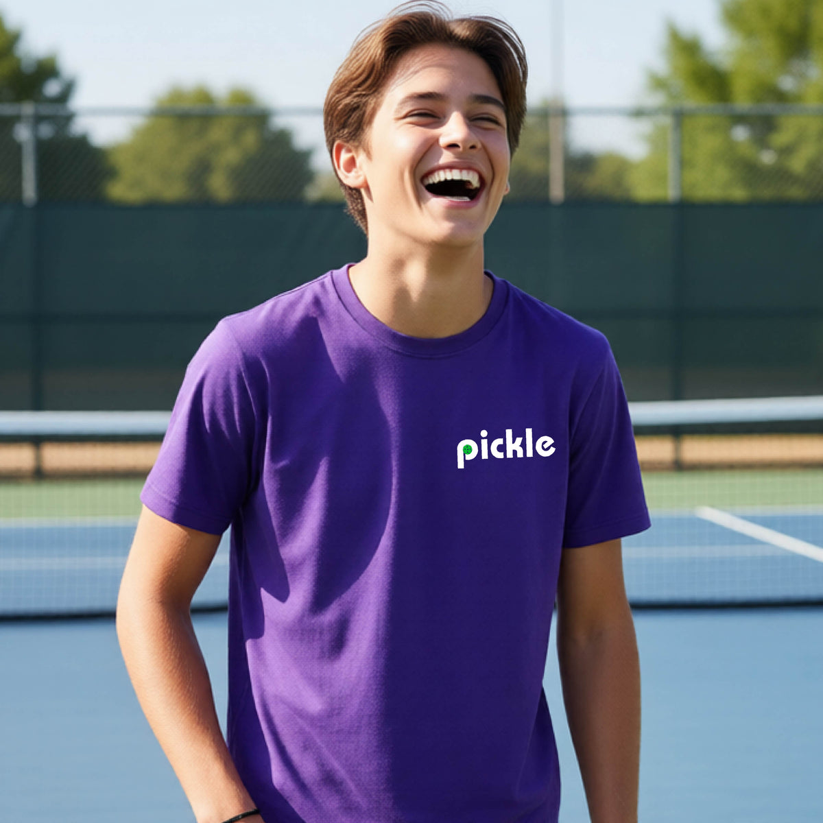 A young person wearing a purple short-sleeve t-shirt with a white graphic of ‘PICKLE’ with a green pickleball integrated into the letter P printed on the front, in front of a pickleball court. 