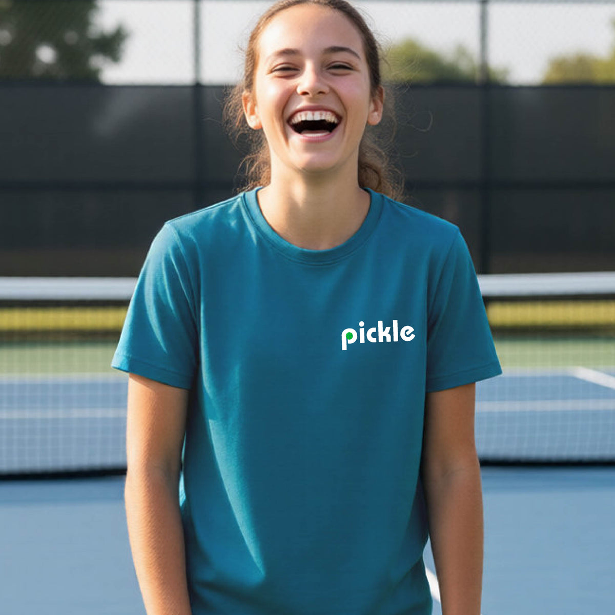 A young person wearing a teal short-sleeve t-shirt with a white graphic of ‘PICKLE’ with a green pickleball integrated into the letter P printed on the front, in front of a pickleball court.