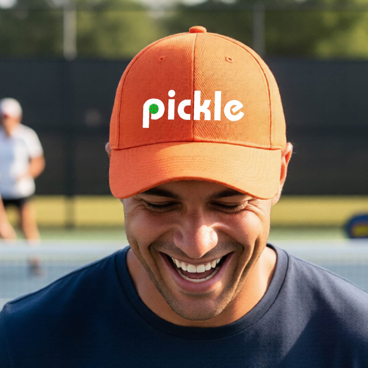 A close-up of an orange pickleball mid-profile hat with a white graphic of ‘PICKLE’ with a green pickleball integrated into the letter P, in front of a pickleball court. 