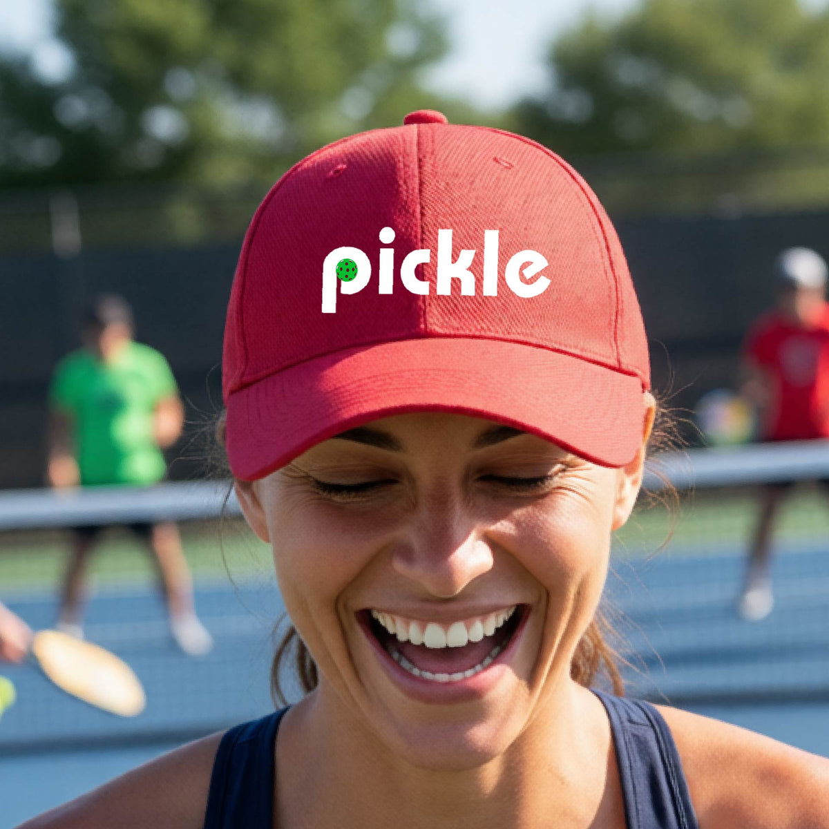 A close-up of a red pickleball mid-profile hat with a white graphic of ‘PICKLE’ with a green pickleball integrated into the letter P, in front of a pickleball court. 