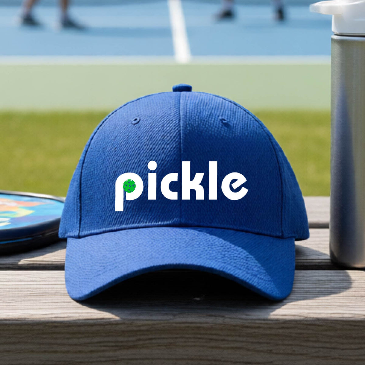 A close-up of a royal blue pickleball mid-profile hat with a white graphic of ‘PICKLE’ with a green pickleball integrated into the letter P, in front of a pickleball court. 