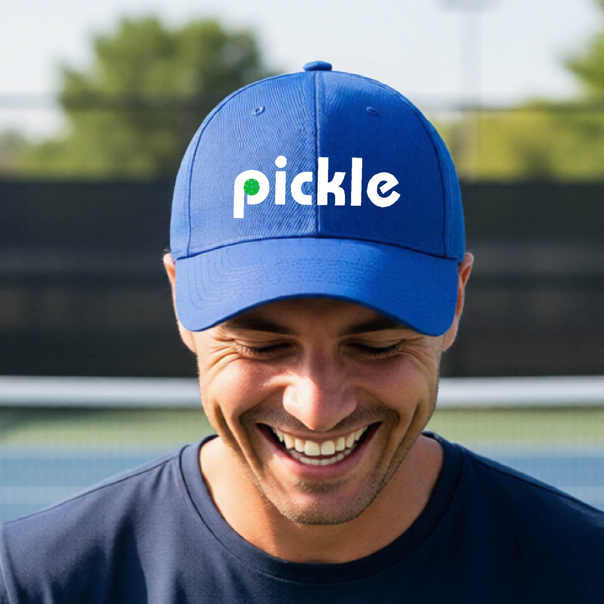 A close-up of a royal blue pickleball mid-profile hat with a white graphic of ‘PICKLE’ with a green pickleball integrated into the letter P, in front of a pickleball court. 
