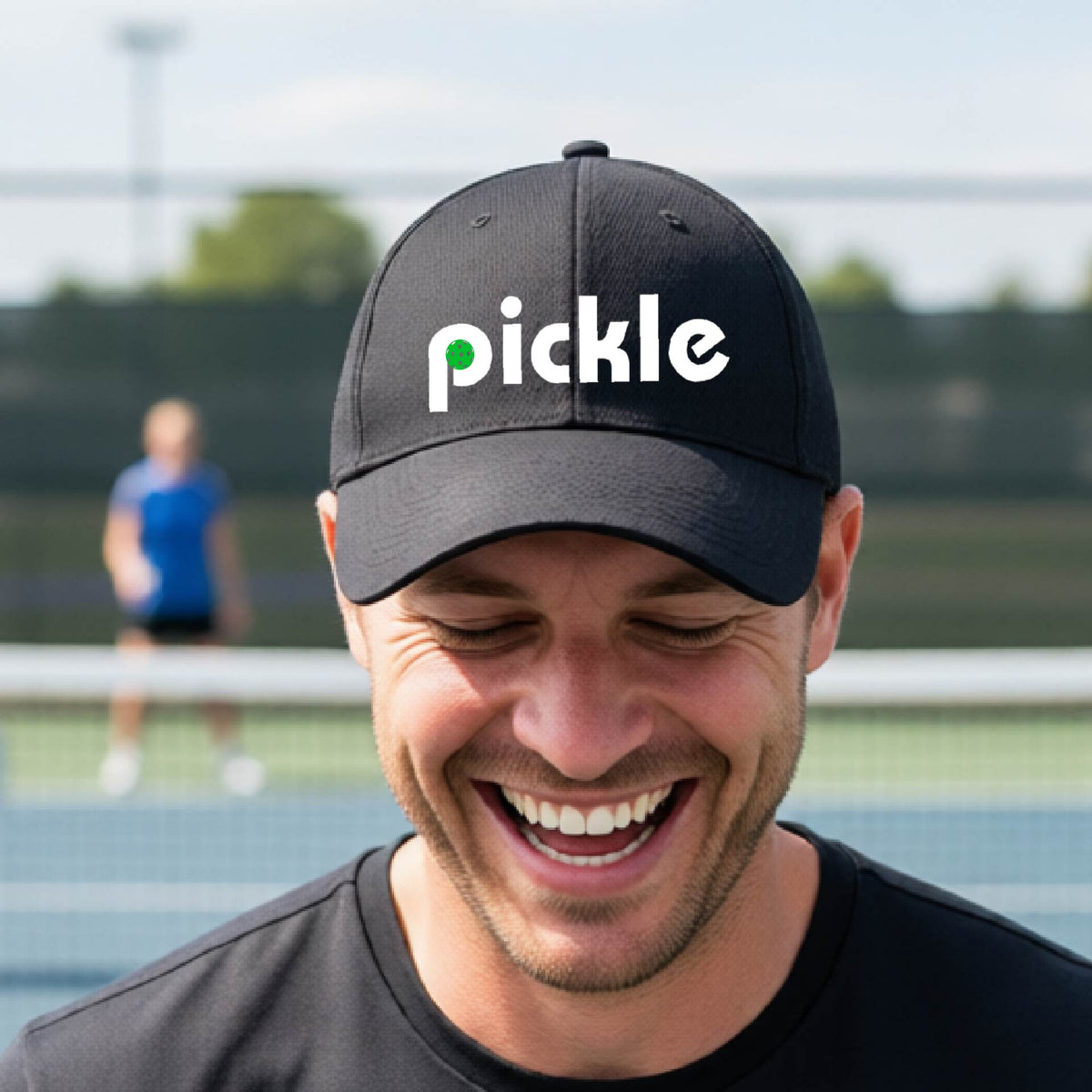 A close-up of a black pickleball mid-profile hat with a white graphic of ‘PICKLE’ with a green pickleball integrated into the letter P, in front of a pickleball court. 
