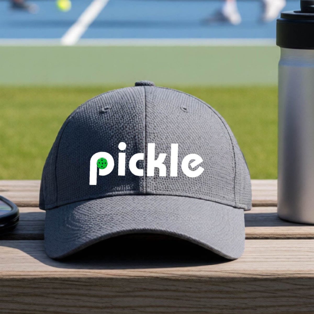 A close-up of a graphite gray pickleball mid-profile hat with a white graphic of ‘PICKLE’ with a green pickleball integrated into the letter P, in front of a pickleball court. 