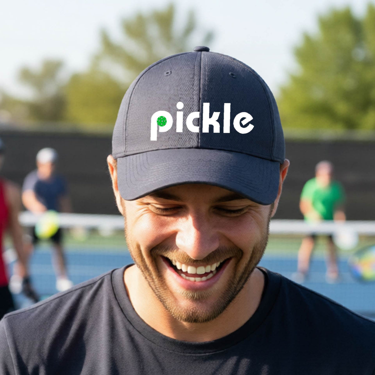 A close-up of a graphite gray pickleball mid-profile hat with a white graphic of ‘PICKLE’ with a green pickleball integrated into the letter P, in front of a pickleball court. 