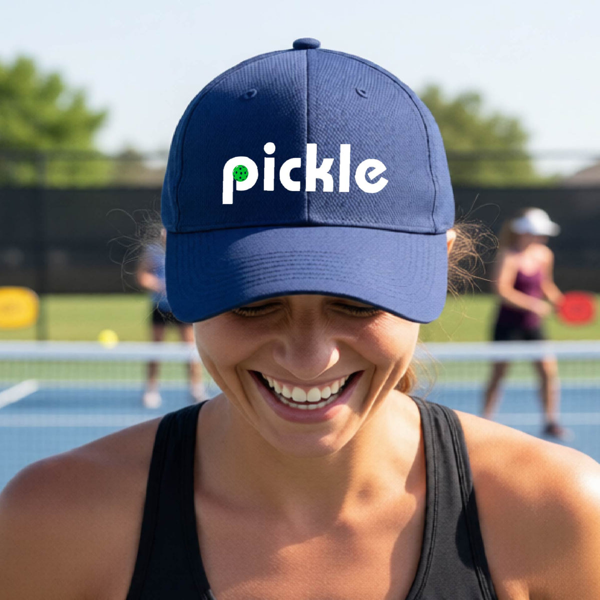 A close-up of a navy blue pickleball mid-profile hat with a white graphic of ‘PICKLE’ with a green pickleball integrated into the letter P, in front of a pickleball court. 