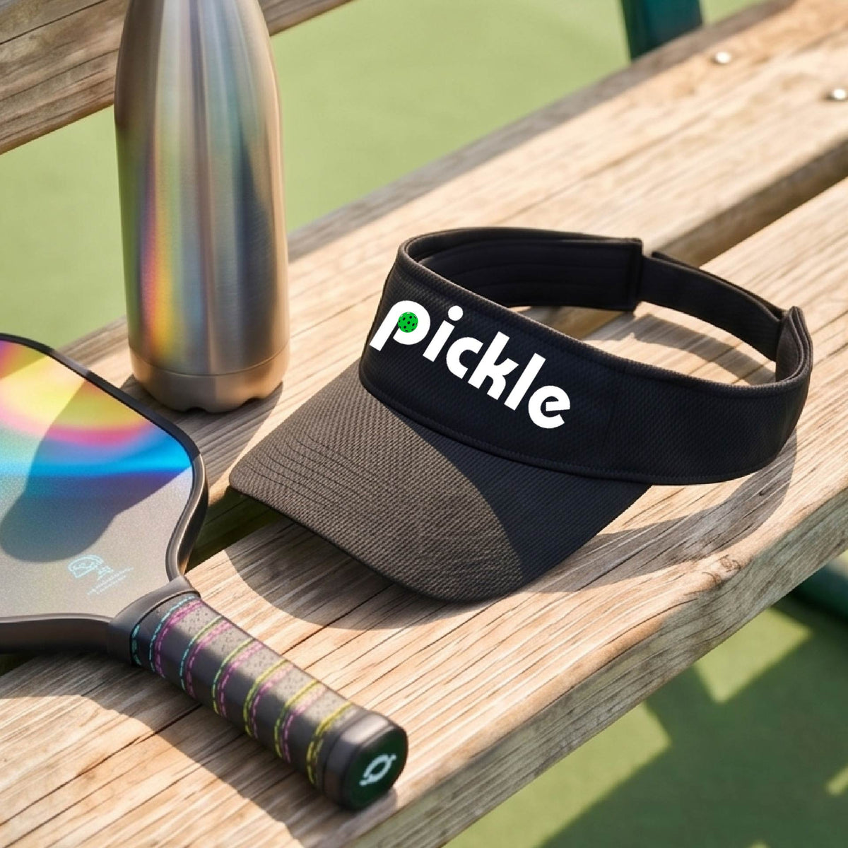 A close-up of a black pickleball visor with a white graphic of ‘PICKLE’ with a green pickleball integrated into the letter P, in front of a pickleball court. 