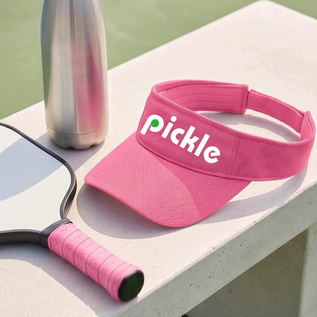 A close-up of a pink pickleball visor with a white graphic of ‘PICKLE’ with a green pickleball integrated into the letter P, in front of a pickleball court. 