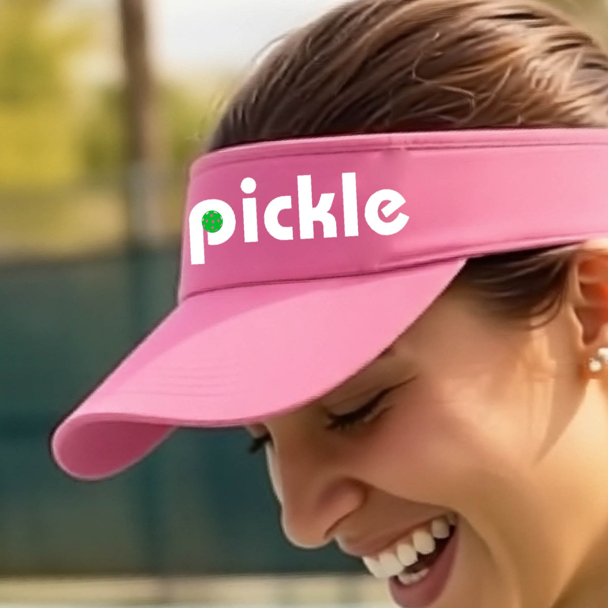 A close-up of a pink pickleball visor with a white graphic of ‘PICKLE’ with a green pickleball integrated into the letter P, in front of a pickleball court. 