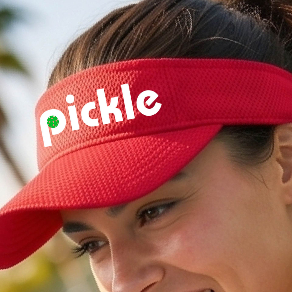 A close-up of a red pickleball visor with a white graphic of ‘PICKLE’ with a green pickleball integrated into the letter P, in front of a pickleball court. 