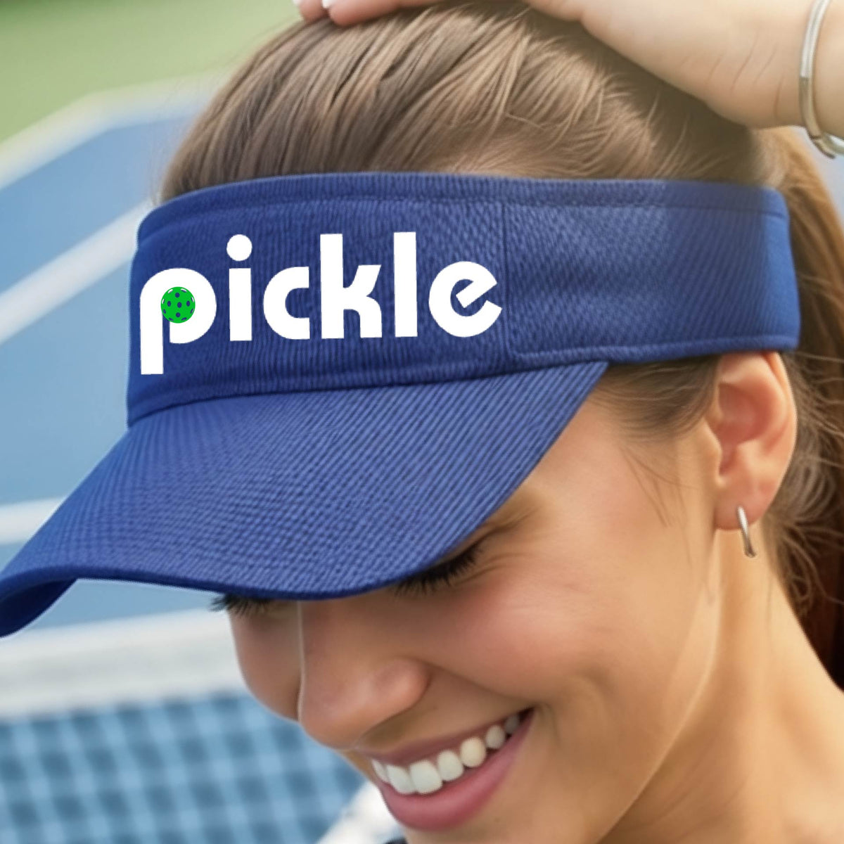 A close-up of a royal blue pickleball visor with a white graphic of ‘PICKLE’ with a green pickleball integrated into the letter P, in front of a pickleball court. 