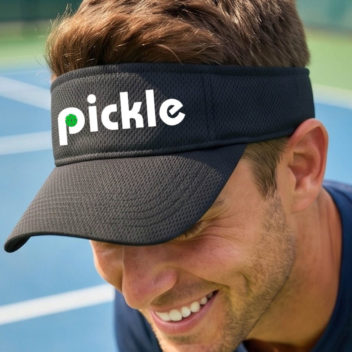 A close-up of a black picklebll visor with a white graphic of ‘PICKLE’ with a green pickleball integrated into the letter P, in front of a pickleball court. 