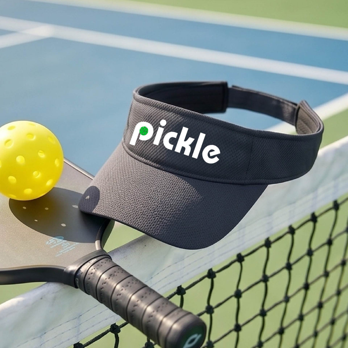 A close-up of a gray pickleball visor with a white graphic of ‘PICKLE’ with a green pickleball integrated into the letter P, in front of a pickleball court. 