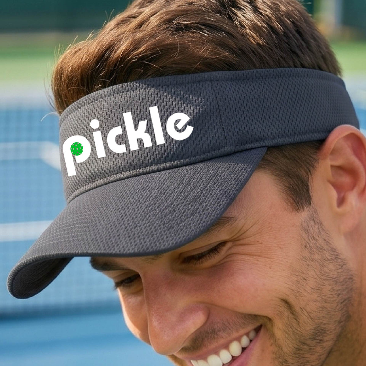 A close-up of a gray pickleball visor with a white graphic of ‘PICKLE’ with a green pickleball integrated into the letter P, in front of a pickleball court. 
