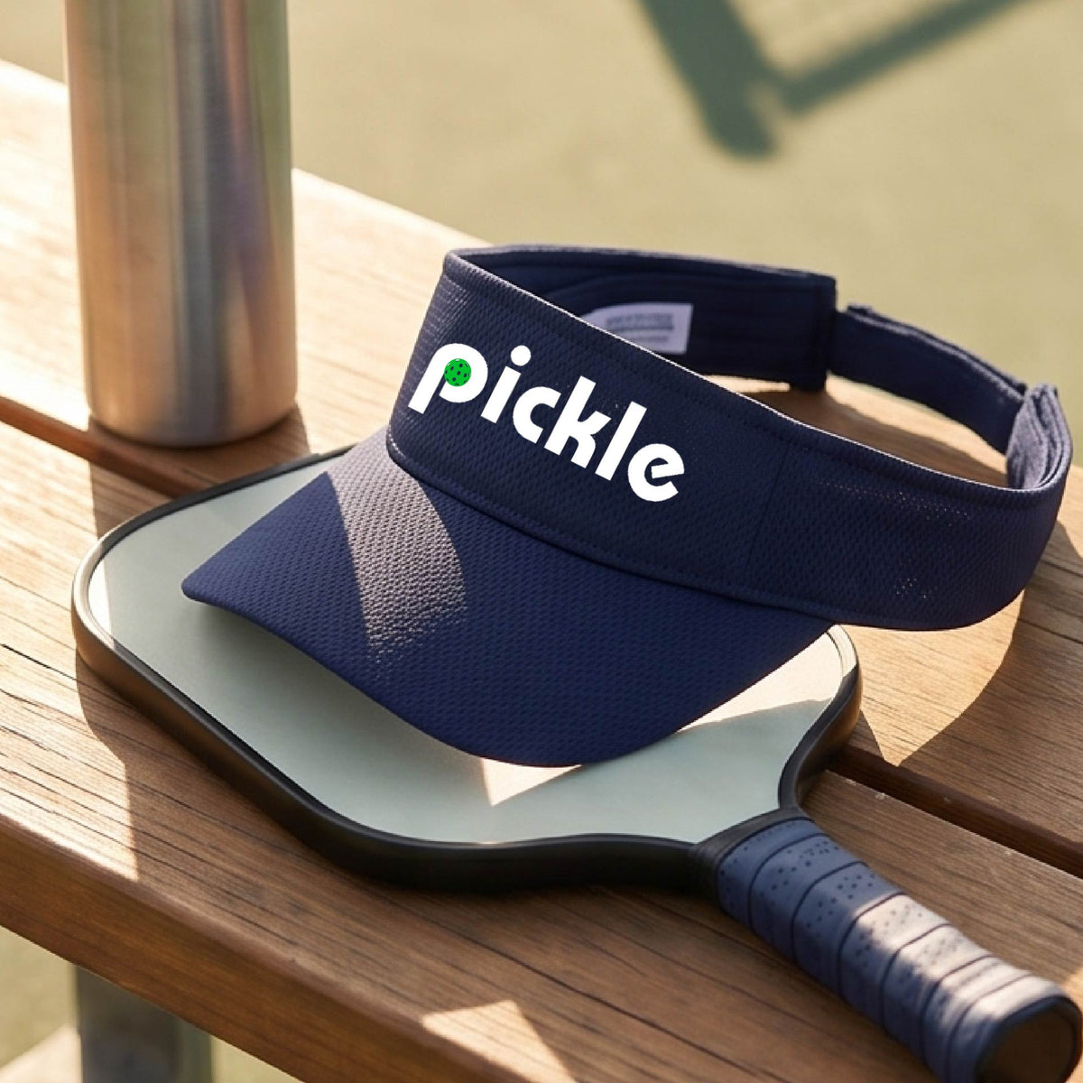 A close-up of a navy blue pickleball visor with a white graphic of ‘PICKLE’ with a green pickleball integrated into the letter P, in front of a pickleball court. 