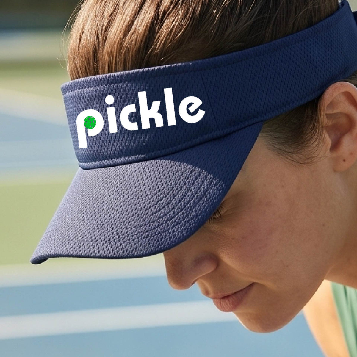 A close-up of a navy blue pickleball visor with a white graphic of ‘PICKLE’ with a green pickleball integrated into the letter P, in front of a pickleball court. 