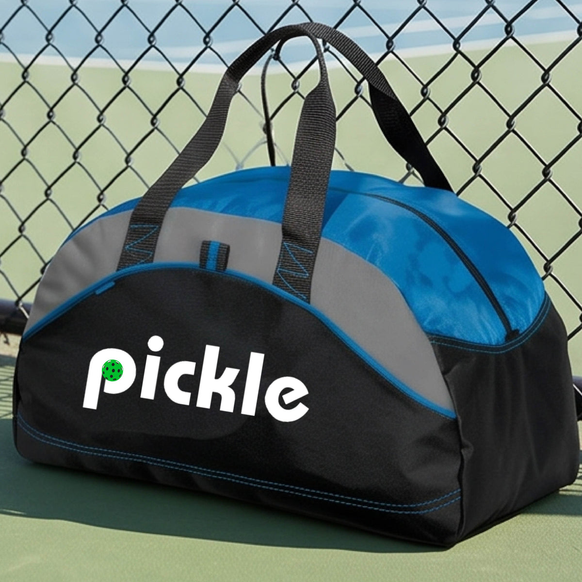 A royal blue, black, and gray duffel bag with the white text  'pickle' printed in white on the front with a green pickleball in the center of hte letter P, placed on a outdoor pickleball court.